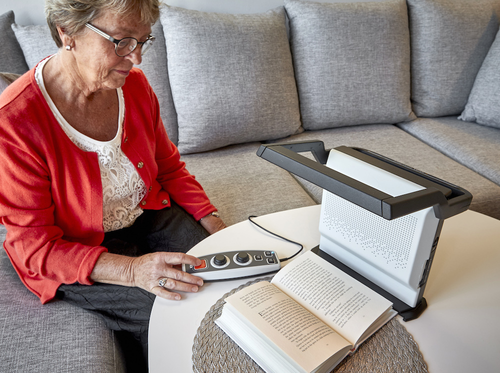 A woman sitting at a table utilizing the LVI Magnilink Voice to read a book.